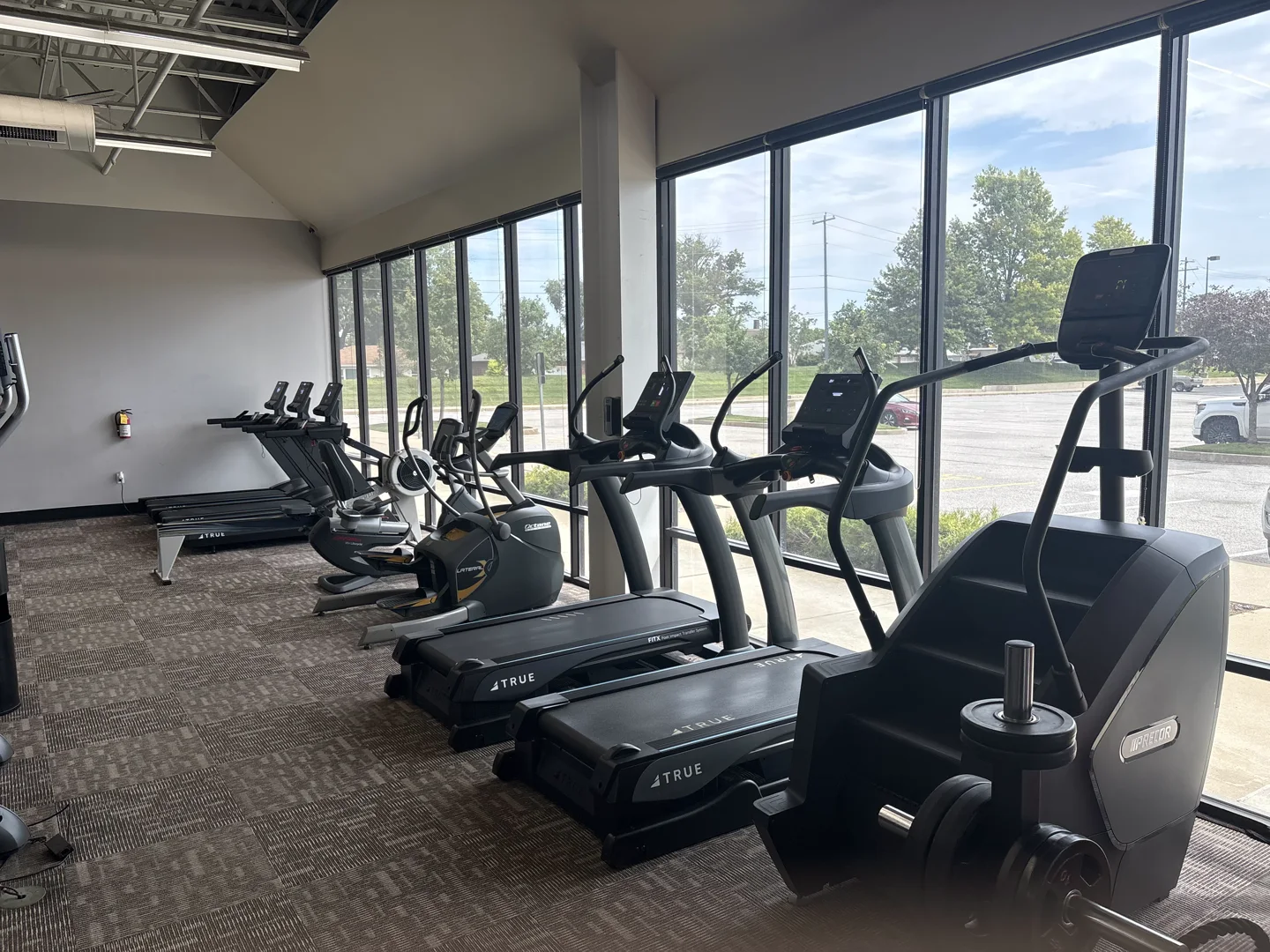 Cardio line at E’ville Fitness with treadmills, stair climber, and cross trainers by the windows